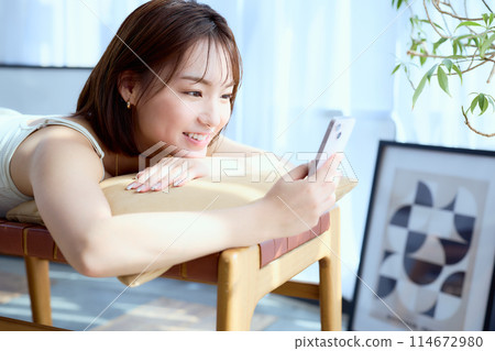 A woman lying on a chair in a room and looking at her smartphone 114672980