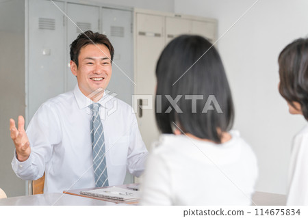 A male teacher, a female student, and her mother having a three-way conference in a school classroom A male teacher, a female student, and her mother having a three-way conference in a school classroom 114675834