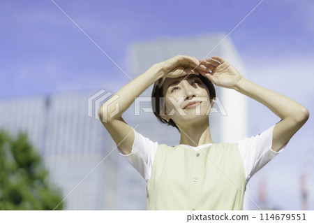 A middle-aged woman holding her hands up in the sunlight in an office district 114679551