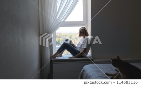 View of a young blond woman sitting by the window with a cup of hot tea. A woman drinks tea while sitting on the windowsill. A woman in a white T-shirt admires the view from the window. 114683110