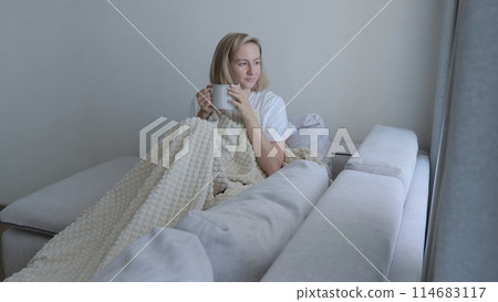A young woman drinks hot tea while lying on the sofa. A woman on the sofa, covered with a blanket, drinks a hot drink. A man is trying to warm up due to the lack of heating in the apartment. 114683117