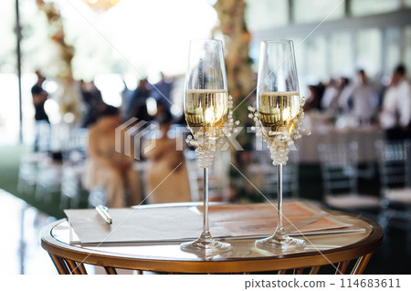 Two wedding glasses stand on a stand against the backdrop of the ceremony arch. 114683611