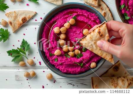 Dipping pita into vibrant beet hummus in bowl with chickpeas, on white rustic wood 114688112