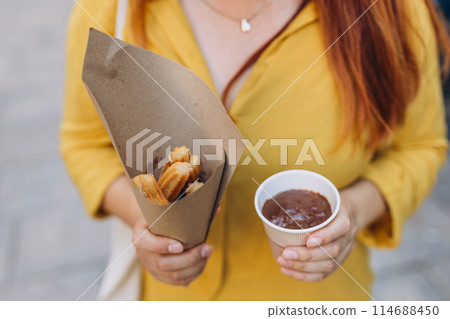 Woman eating traditional Churros, a fried pastry with chocolate on a city street. Food concept Woman eating traditional Churros, a fried pastry with chocolate on a city street. Food concept 114688450