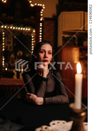 Vertical portrait of enigmatic fortune teller in black robe sitting at table in dark esoteric room by light of burning candles, looking at camera with serious expression. Concept of divination, magic. 114696062