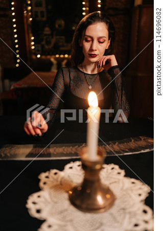 Mysterious fortune teller in black robe sitting at table by light of burning candles, looking down with serious pensive expression, in dark esoteric room with mystical atmosphere. Concept divination 114696082