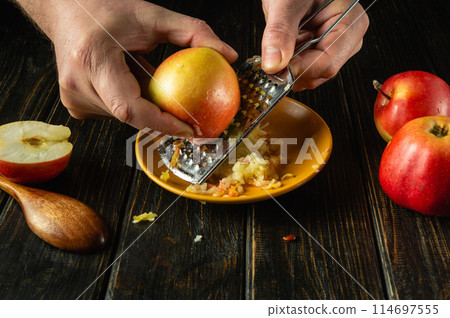 The hands of a cook with a grater grate a raw apple on the kitchen table. Low key concept of preparing a healthy fruit breakfast The hands of a cook with a grater grate a raw apple on the kitchen table. Low key concept of preparing a healthy fruit breakfast 114697555