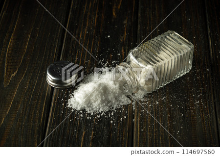 A pile of salt is poured from a hodgepodge on a black table. The concept of excessive salt consumption, which can be the cause of various diseases. Place for advertising. A pile of salt is poured from a hodgepodge on a black table. The concept of excessive salt consumption, which can be the cause of various diseases. Place for advertising. 114697560