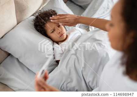 A concerned young African American mother is gently placing her hand on her child forehead to check for a fever while the child lies in bed. The mother appears attentive and caring 114698947