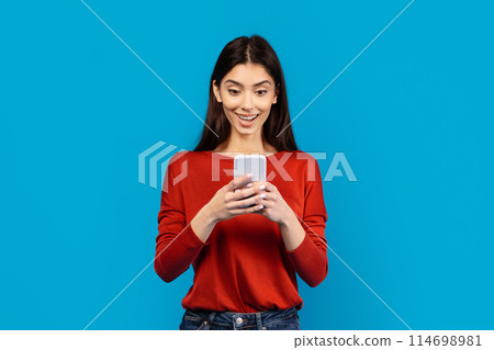 A woman wearing a red shirt is holding a cell phone in her hand, looking at the screen. She appears focused on whatever is displayed on the device, possibly texting or browsing online. 114698981
