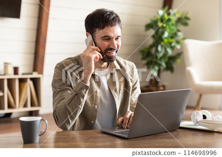 A man is multitasking by talking on a cell phone and using a laptop simultaneously. He appears focused on his work, balancing communication and productivity on two devices. 114698996