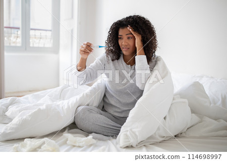 A young Hispanic woman wrapped in a white blanket is sitting up in bed with a concerned expression as she looks at a thermometer. 114698997