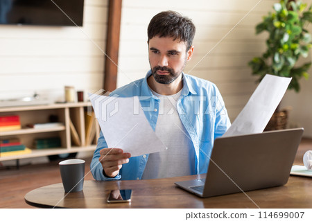A man sits in front of his laptop, holding a piece of paper in his hand. He appears focused and engaged as he works on his computer, checking report 114699007