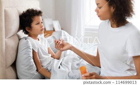 African American mother is gently holding her child hand and checking his temperature while he rests in bed, appearing unwell 114699013