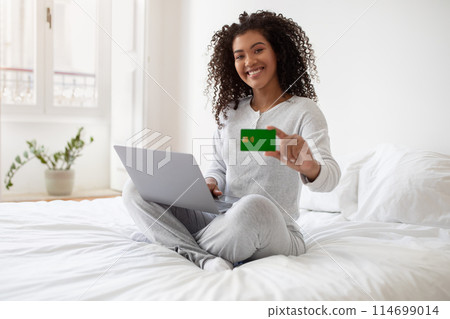 A Hispanic woman is seated on a bed, holding a laptop in one hand and a credit card in the other. She seems focused on the screen, possibly making an online purchase or managing finances. 114699014