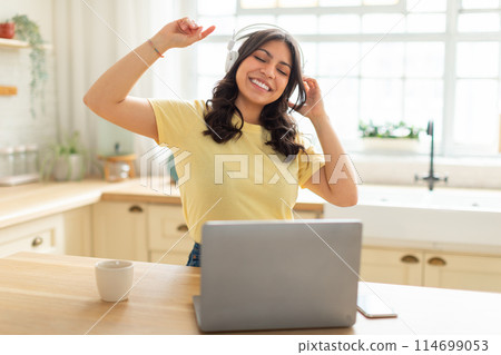 A cheerful young middle eastern woman is seen with closed eyes and headphones on, dancing slightly with her arms raised in joy as she listens to music on laptop, kitchen interior A cheerful young middle eastern woman is seen with closed eyes and headphones on, dancing slightly with her arms raised in joy as she listens to music on laptop, kitchen interior 114699053