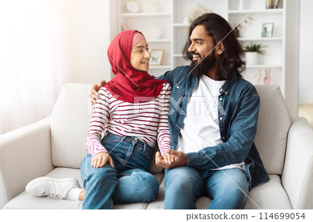 Loving eastern man and a woman are seated together on a couch in a room. They appear to be engaged in conversation or watching television Loving eastern man and a woman are seated together on a couch in a room. They appear to be engaged in conversation or watching television 114699054