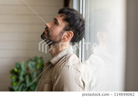 A man standing indoors beside a window, gazing outside at the scenery. The man appears contemplative as he observes the outdoor surroundings through the clear glass pane. 114699062