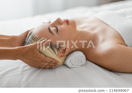A woman is lying down on a massage table at a spa while a professional masseuse is giving her a head massage. The woman appears to be relaxed and enjoying the soothing touch on her scalp. 114699067