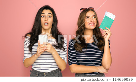 Two young women stand against a vivid pink backdrop, one holding her smartphone with a surprised expression while the other, holding a pair of travel tickets, perhaps planning a journey. Two young women stand against a vivid pink backdrop, one holding her smartphone with a surprised expression while the other, holding a pair of travel tickets, perhaps planning a journey. 114699089