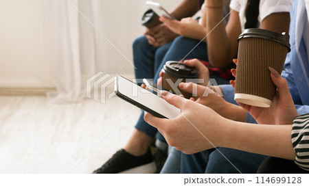 A group of teenagers are seated closely together, with their focus on their handheld smartphones. One individual is holding a paper coffee cup, suggesting a relaxed, social setting, cropped 114699128