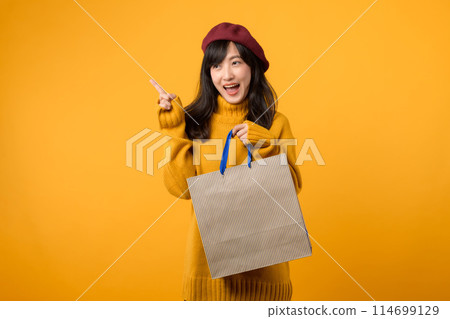 Young Asian woman in her 30s, wearing a yellow sweater and red beret, indulges in a shopping extravaganza and pointing finger to free copy space against a vibrant yellow backdrop. 114699129