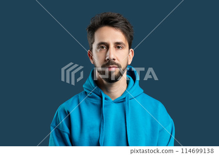 A man with a full beard is standing while wearing a blue hoodie. He appears relaxed and casual, with his hands in his pockets. The background is unremarkable, allowing the man to stand out. A man with a full beard is standing while wearing a blue hoodie. He appears relaxed and casual, with his hands in his pockets. The background is unremarkable, allowing the man to stand out. 114699138