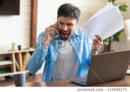 A visibly upset man in a blue shirt talks on his mobile phone, with one hand holding papers and the other gesturing angrily. He sits at a wooden table facing a laptop A visibly upset man in a blue shirt talks on his mobile phone, with one hand holding papers and the other gesturing angrily. He sits at a wooden table facing a laptop 114699173