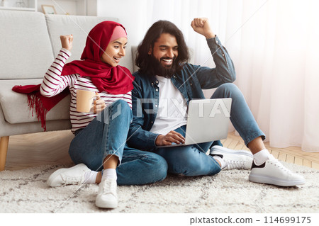 Indian man and woman are seated on the floor, working on a laptop together. They appear focused and engaged in their work, surrounded by a simple, neutral-colored room. 114699175