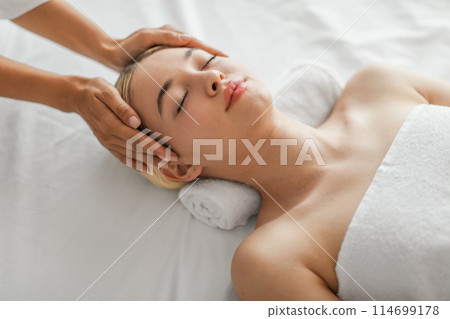 A serene young woman is lying down, eyes closed, experiencing the soothing touch of a head massage at a spa. A pair of professional hands gently works to relieve tension 114699178