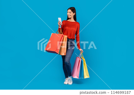 A woman holding shopping bags in one hand and a cell phone in the other. She appears focused on her phone, possibly checking messages or making calls while multitasking with her shopping. 114699187