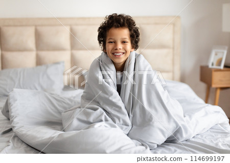 African American child is sitting on a bed, wrapped snugly in a soft blanket. The child eyes are closed, indicating sleep or rest. The room is dimly lit, creating a cozy atmosphere. 114699197