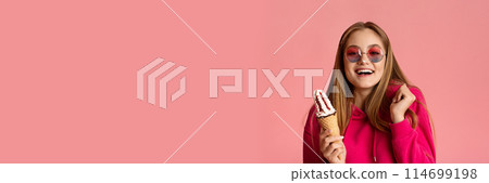 Smiling teenage girl in round glasses with ice cream in her hands gesturing and grimacing on pink studio background, panorama with copy space 114699198