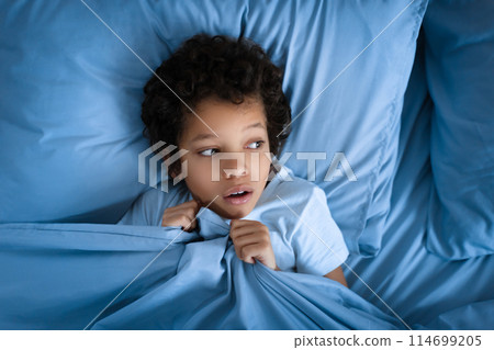 Scared African American young boy with curly hair lies awake in a blue bed during night, clutching the bedding close to his chin with a look of fright 114699205