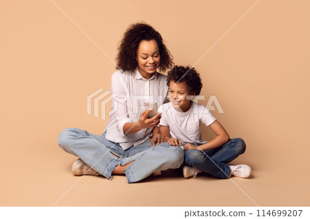 African American woman and a child are seated on the floor, both looking intently at phone screen. The woman appears engaged in conversation or viewing content, while the child watches attentively. 114699207