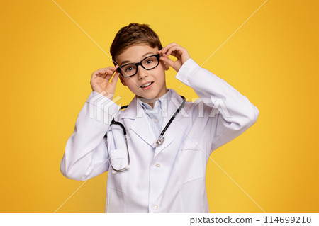 Pharmacy occupation. Friendly teen boy in doctor uniform posing, orange studio background 114699210