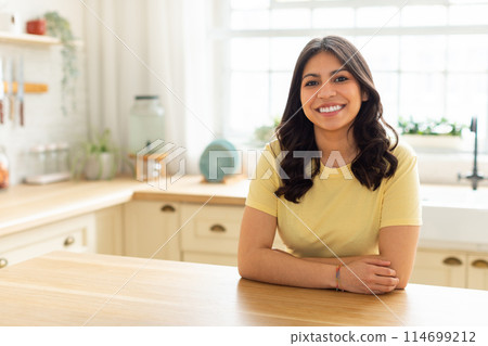 A middle eastern woman is sitting at a wooden table in a white kitchen. The kitchen is bright with natural light coming in from a window. 114699212
