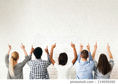 A group of friends stands in front of a blank white wall, all pointing upwards. Their gestures suggest they are indicating or drawing attention to something above them, back view 114699260
