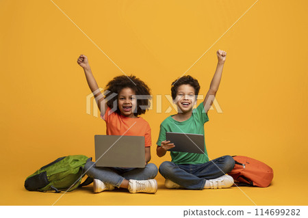 Two African American children, a boy and a girl, are sitting on the floor with laptop and digital tablet in front of them, raising hands up, yellow background 114699282