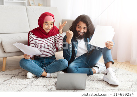 A young Eastern man and woman sit on the floor beside a gray couch, engaged with papers and a laptop in a well-lit room. Both are smiling broadly 114699284