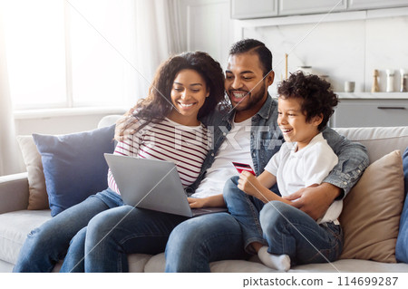 African American man, woman, and child are seated on a sofa. They are all looking at a laptop screen, engaged in an activity. The room is well-lit and cozy, creating a warm atmosphere. African American man, woman, and child are seated on a sofa. They are all looking at a laptop screen, engaged in an activity. The room is well-lit and cozy, creating a warm atmosphere. 114699287