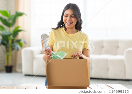 A cheerful middle eastern woman is standing in a brightly lit living room, looking delighted as she unpacks modern iron from a cardboard box. A cheerful middle eastern woman is standing in a brightly lit living room, looking delighted as she unpacks modern iron from a cardboard box. 114699315
