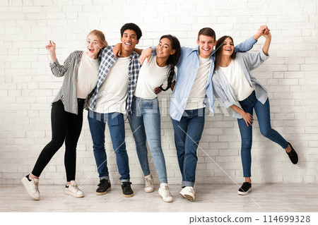 A group of five multiethnic teenagers are standing in a row against a white brick wall, smiling and laughing. They are dressed in casual clothing 114699328