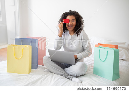 A Hispanic woman wearing casual clothes is sitting on a bed surrounded by colorful shopping bags and a laptop, cover one of her eyes with credit card 114699370