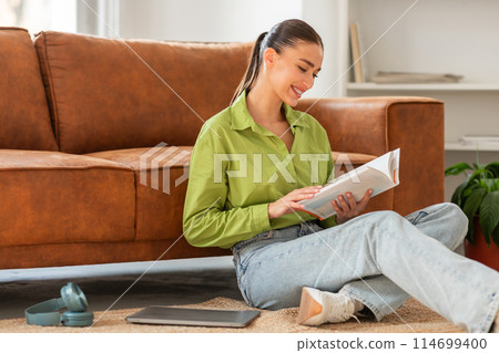 A woman is seated on the floor, engrossed in a book she is reading. The woman appears focused as she flips through the pages, immersed in the content of the book. 114699400
