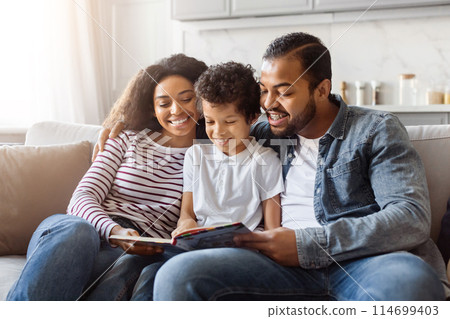 Black family consisting of parents and children are seated together on a couch, engrossed in reading a book. The parents and children are focused on the pages, expressing interest and engagement. Black family consisting of parents and children are seated together on a couch, engrossed in reading a book. The parents and children are focused on the pages, expressing interest and engagement. 114699403