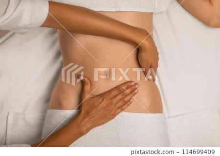 A physical therapists hands are assisting a patient by guiding and supporting their abdomen during rehabilitation exercises. The patient is lying down, wearing a white top and partially visible pants A physical therapists hands are assisting a patient by guiding and supporting their abdomen during rehabilitation exercises. The patient is lying down, wearing a white top and partially visible pants 114699409