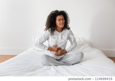 A young Hispanic woman with curly hair sits cross-legged on a bed, clutching her stomach with a pained expression on her face suggesting discomfort or illness 114699419