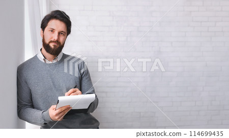 Waiting for patient. Friendly psychologist standing with clipboard, leaning at wall in office, empty space 114699435