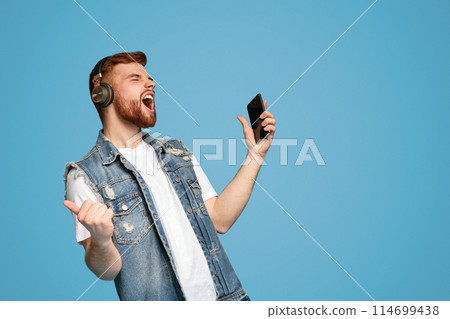 Cool young bearded man singing in headphones and enjoying music with closed eyes on blue background, copy space Cool young bearded man singing in headphones and enjoying music with closed eyes on blue background, copy space 114699438